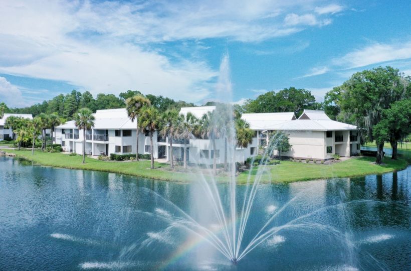 The image depicts white buildings adjacent to a serene body of water, with a large fountain in the foreground and lush greenery in the background.