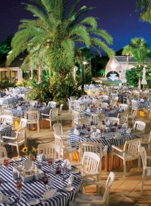 Outdoor evening event setup with tables, chairs, and a pool under palm trees.