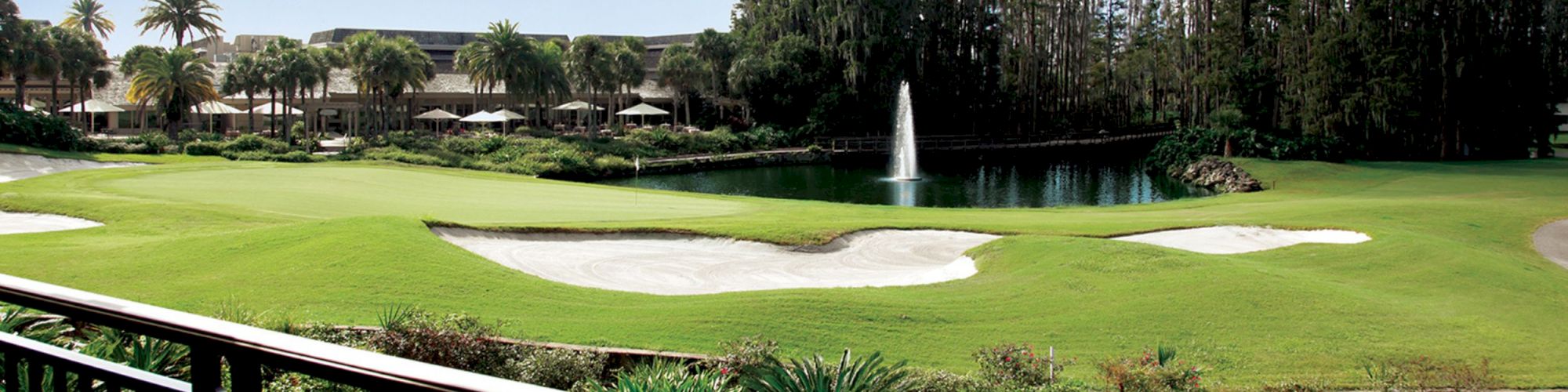 A well-manicured golf course featuring sand traps, a water hazard with a fountain, and a clubhouse surrounded by palm trees and lush vegetation.