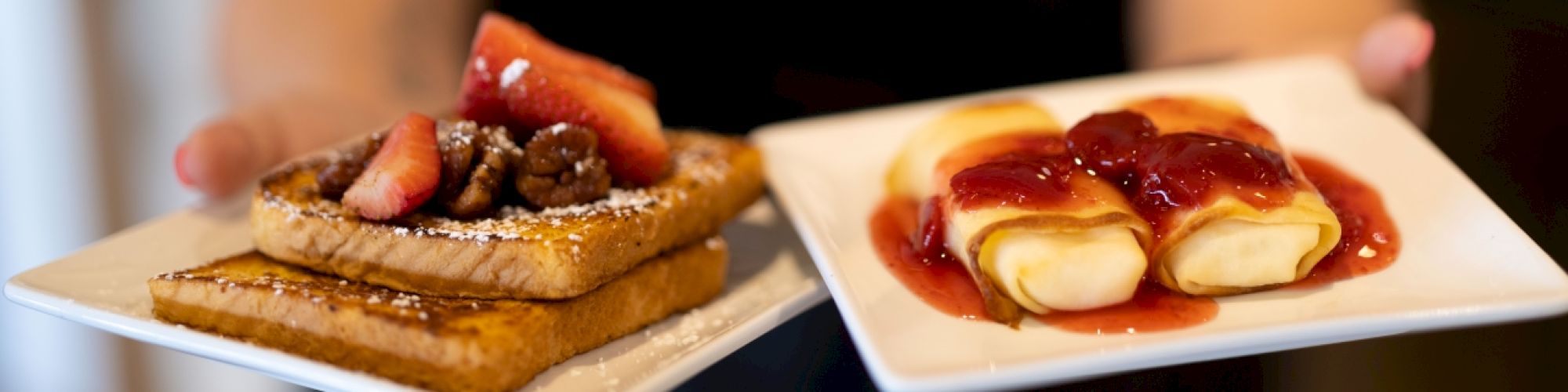 A person is holding two plates of food: one with French toast topped with strawberries, and one with crepes covered in a red sauce.