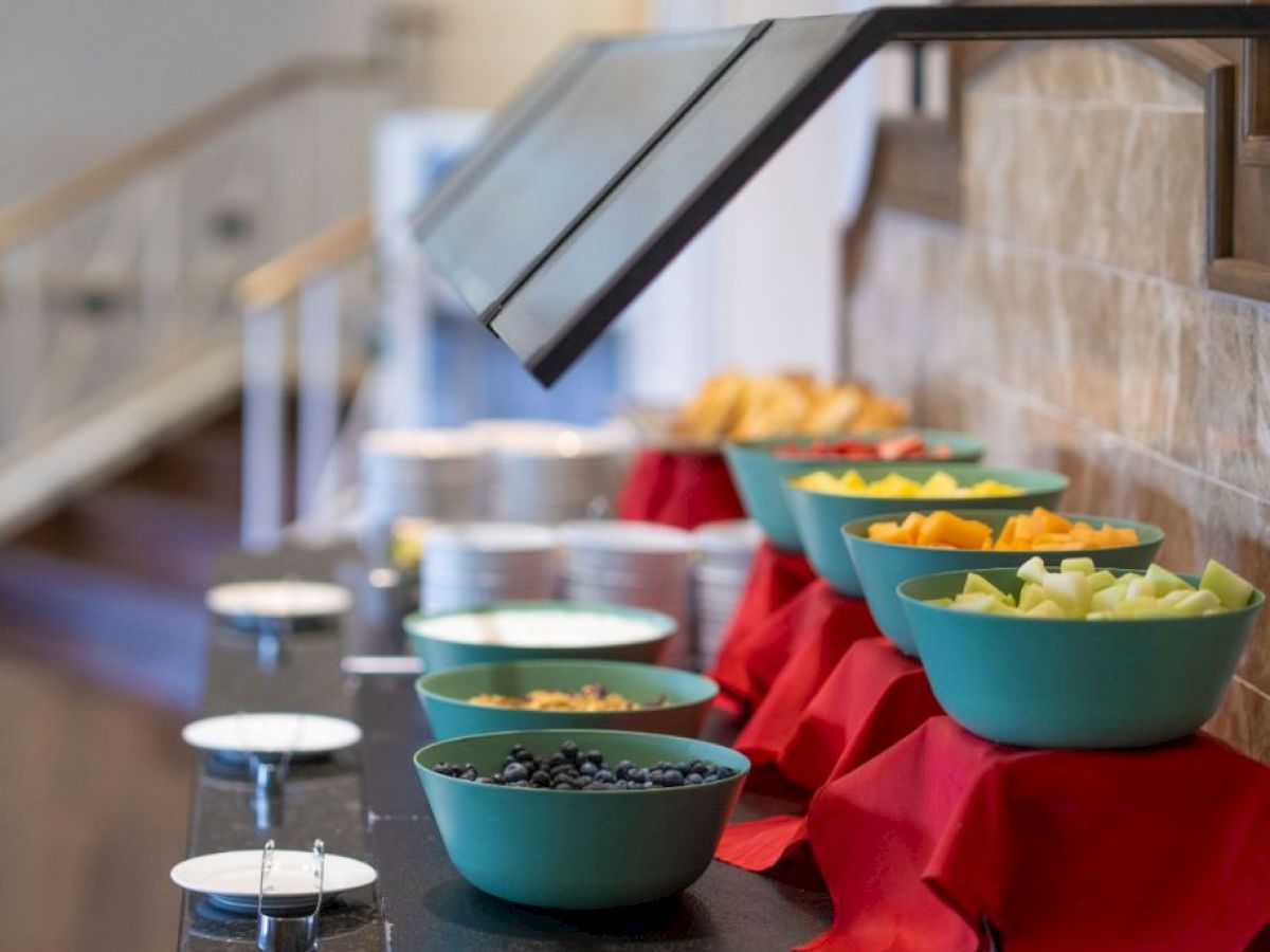 The image shows a buffet setup with bowls of cut fruits on a counter, covered by a sneeze guard, adorned with red napkins underneath the bowls.