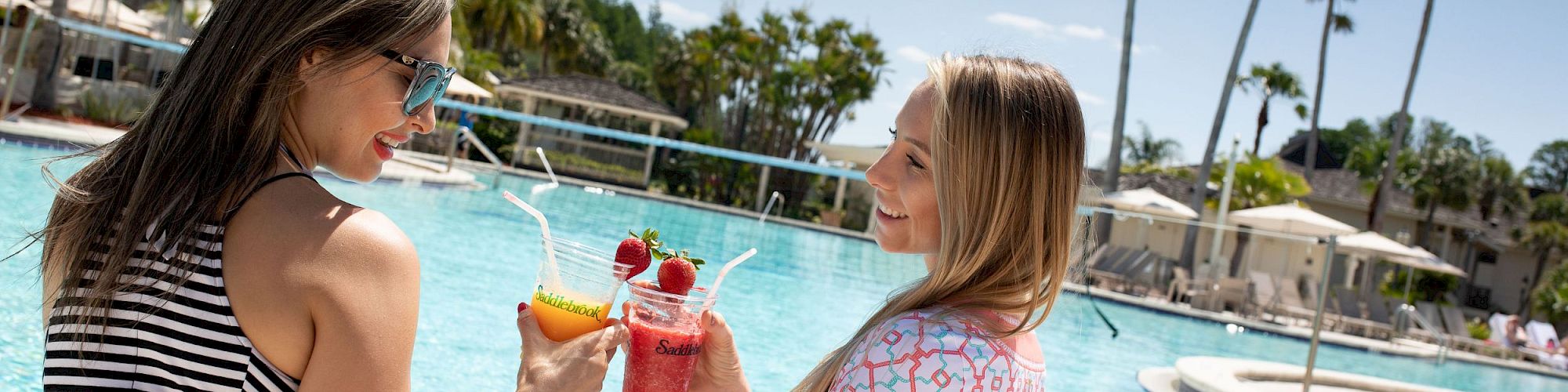 Two women clink their colorful drinks while standing at a sunny outdoor pool, surrounded by palm trees and resort facilities.