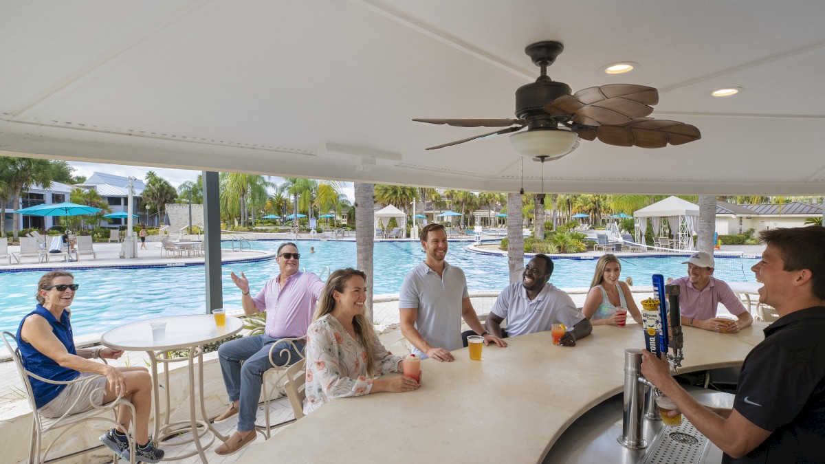 A group of people are enjoying drinks at a poolside bar, engaging with the bartender, with a swimming pool visible in the background.