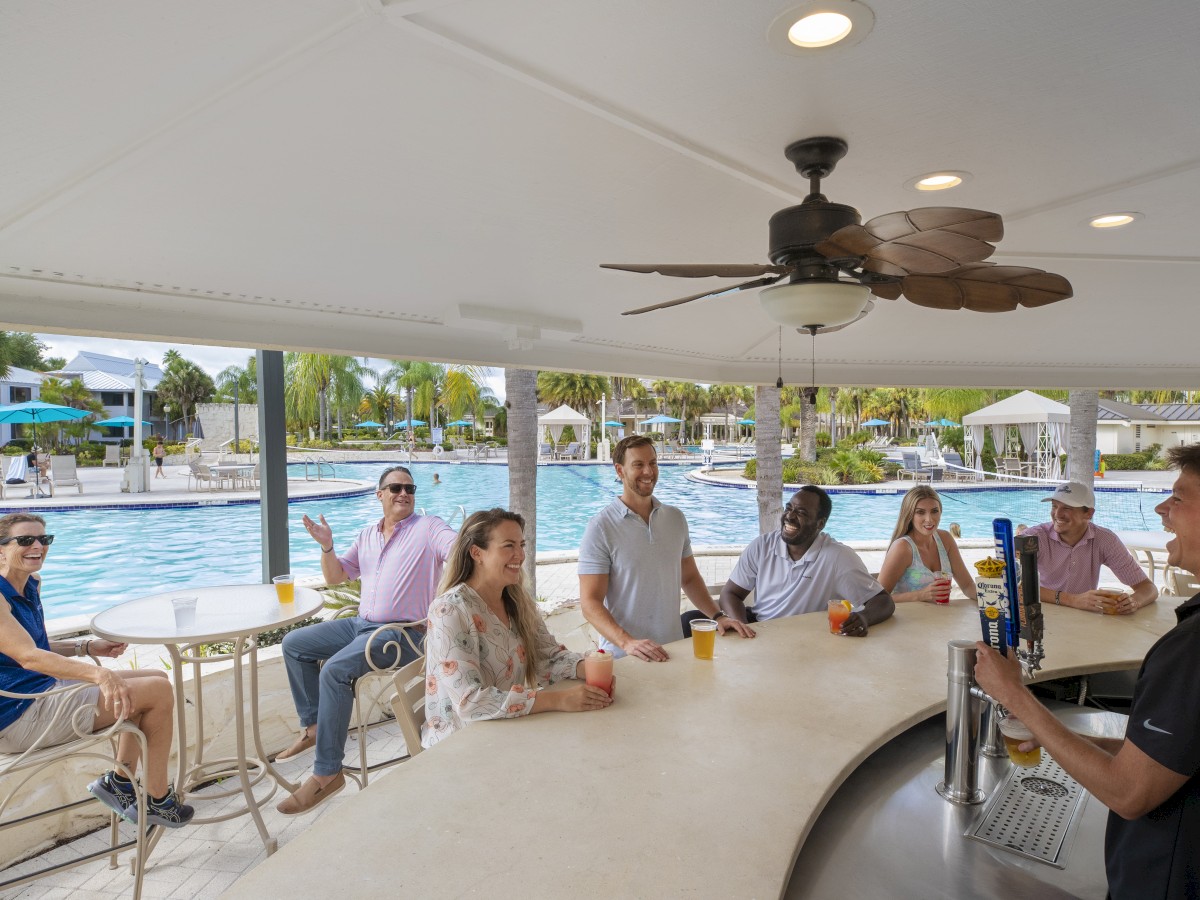 A group of people are enjoying drinks at a poolside bar, engaging with the bartender, with a swimming pool visible in the background.