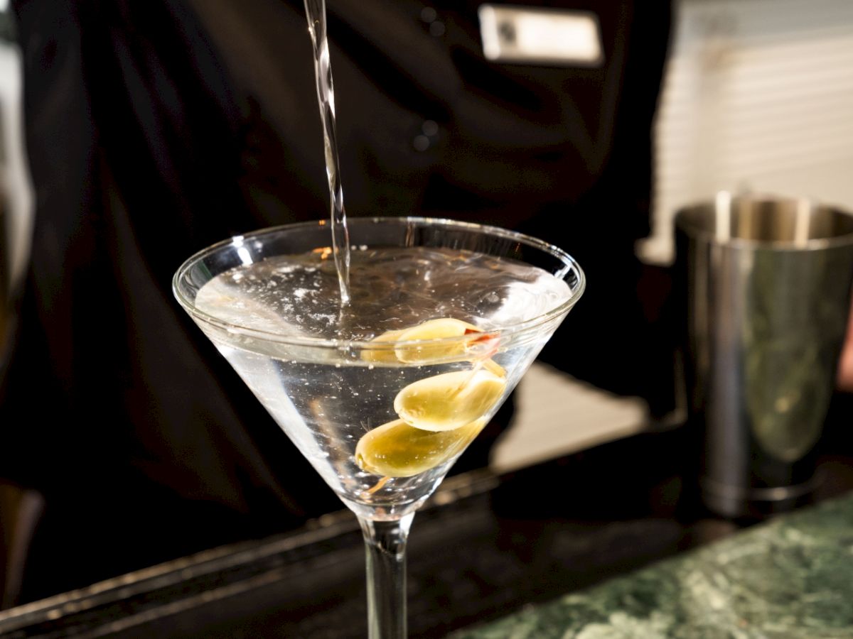 A bartender is pouring a drink into a martini glass with olives on a bar counter. The bartender is wearing a black shirt with a name tag.