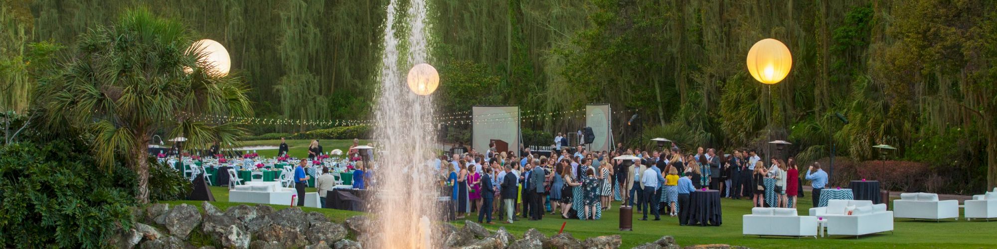 Outdoor event with a fountain, large group of people, string lights, tables, and illuminated orbs in a garden setting.