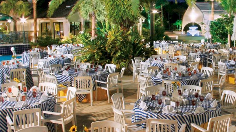 An outdoor event setup with round tables covered in striped tablecloths, surrounded by white chairs, and a palm tree in the center, at night.