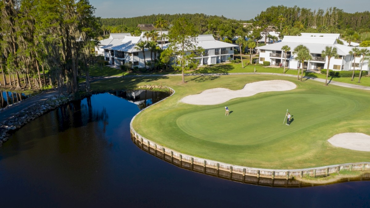 A golf course with a putting green, surrounded by water, sand traps, and residential buildings in the background under a clear sky.