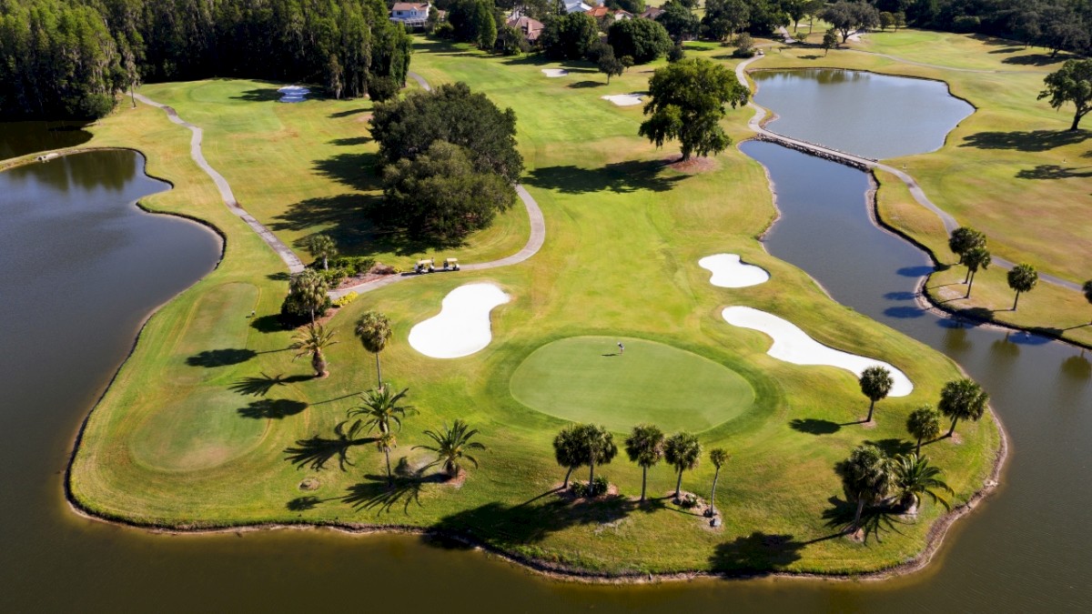 An aerial view of a golf course featuring a green surrounded by water with sand traps, trees, and pathways visible throughout the landscape.