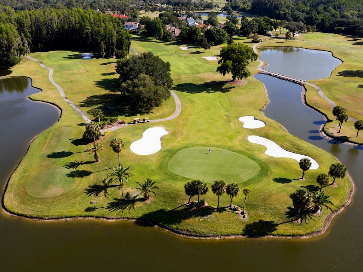 An aerial view of a golf course featuring a green surrounded by water with sand traps, trees, and pathways visible throughout the landscape.
