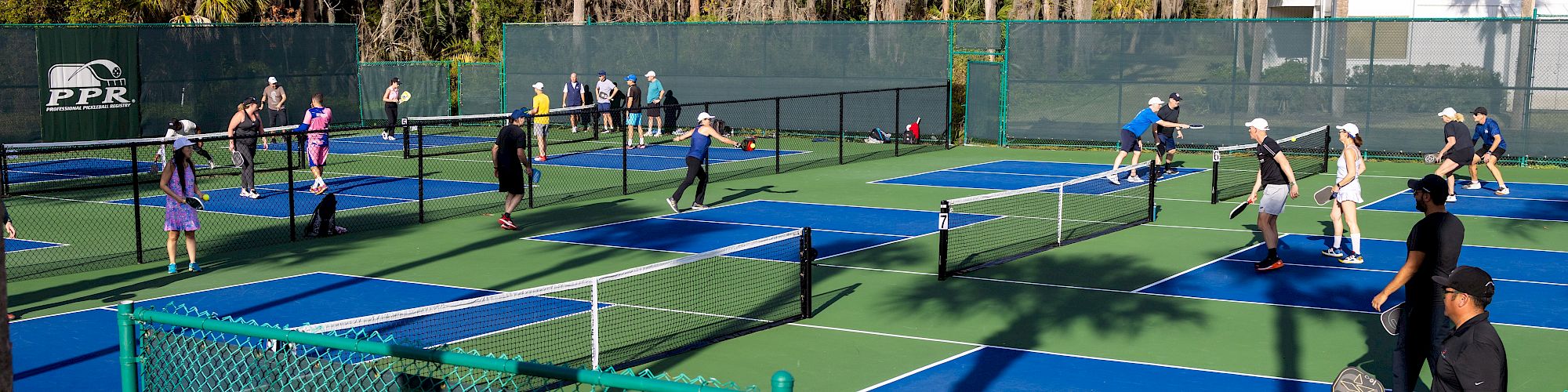 The image shows multiple people playing pickleball on several blue and green courts surrounded by a green fence and trees.
