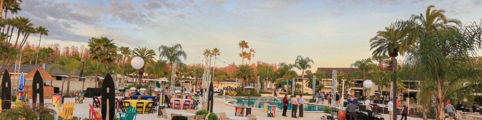An outdoor event with people around a pool, palm trees, decorative lights, and various seating arrangements.