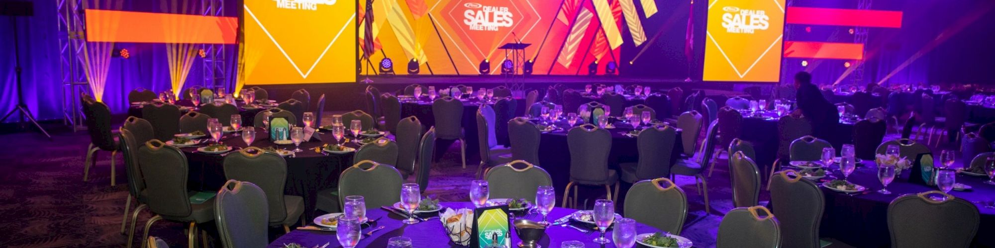 A conference or gala setup with round tables, plates of salad, glasses, and a stage with a large screen showing 