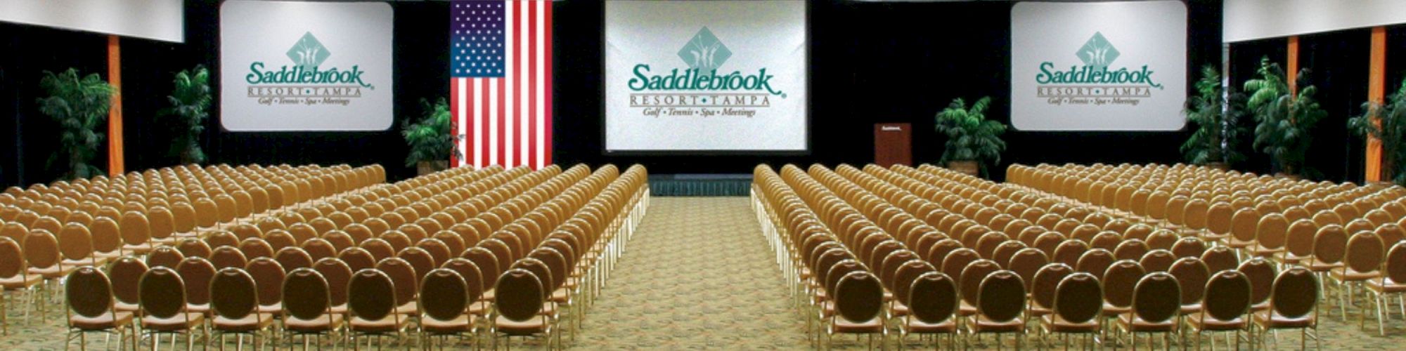 An empty conference room boasts rows of chairs facing a stage with screens, a U.S. flag, and branding for Saddlebrook Resort Tampa.