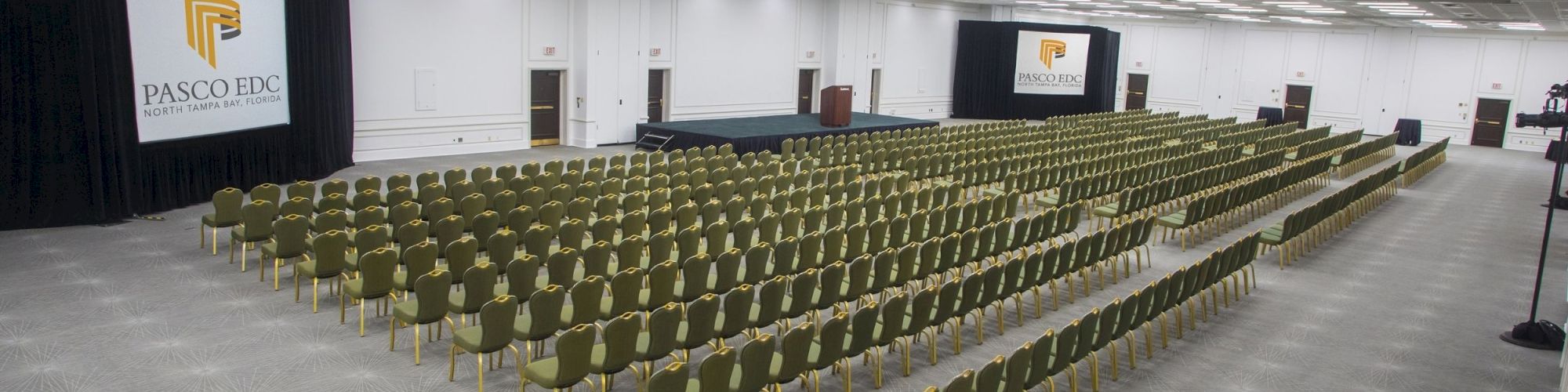 An empty conference room with rows of green chairs facing a stage with two screens displaying 