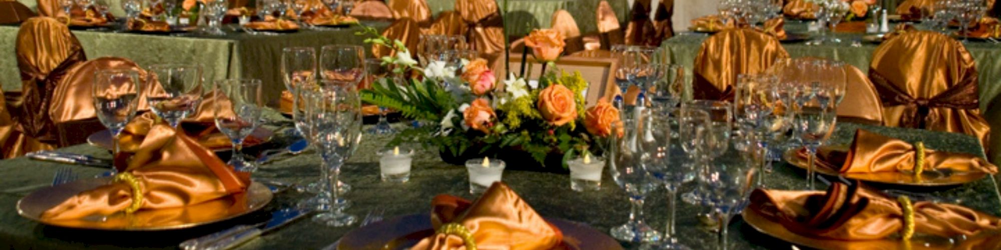 A banquet setup with elegantly decorated tables featuring orange napkins, floral centerpieces, multiple glasses, and dim lighting in the background.