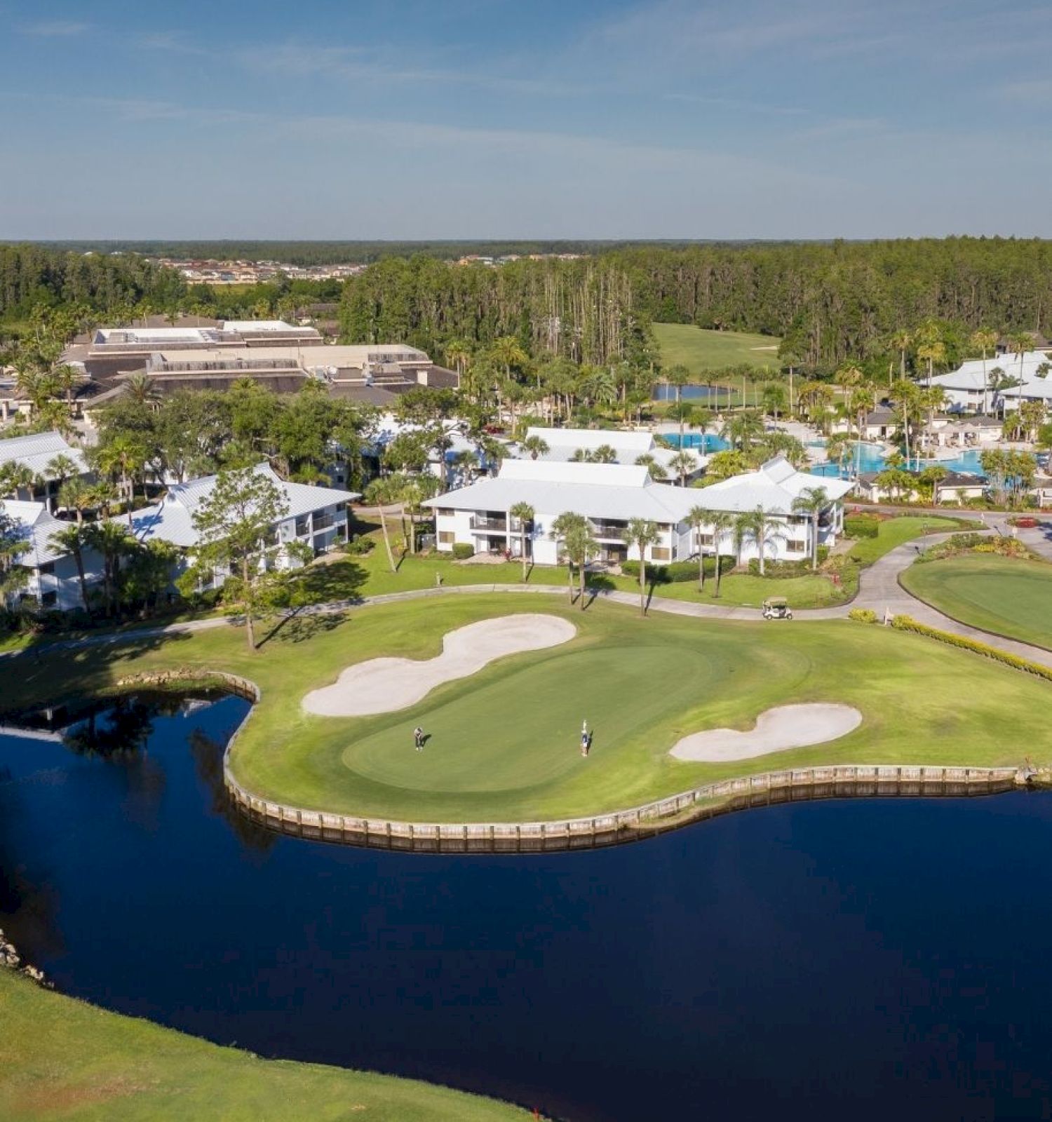 The image shows a scenic aerial view of a golf course with sand traps, a pond, and adjacent buildings surrounded by trees, under a clear sky.