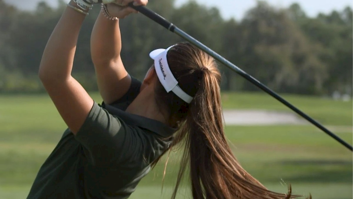 A person with long hair and a visor is swinging a golf club on a green golf course with trees in the background.