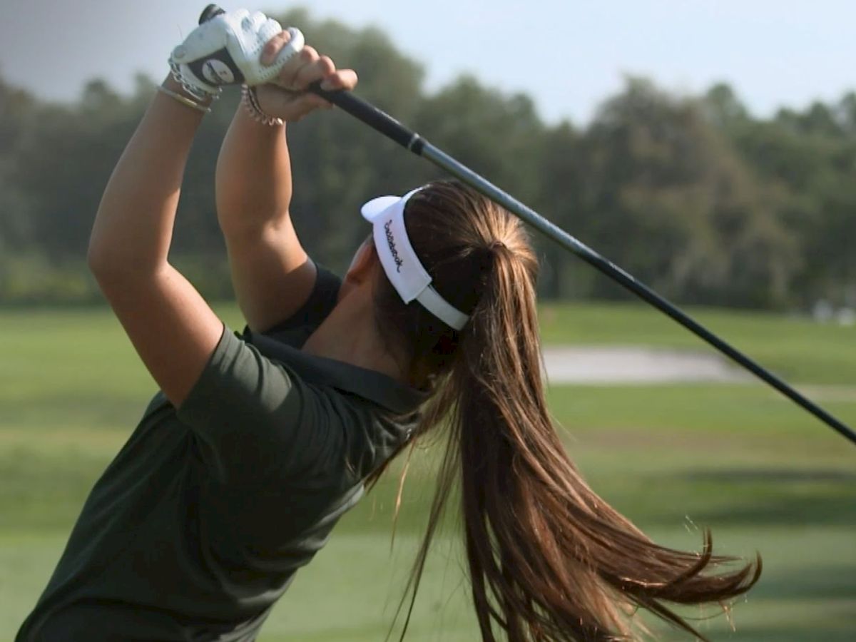 A person with long hair and a visor is swinging a golf club on a green golf course with trees in the background.