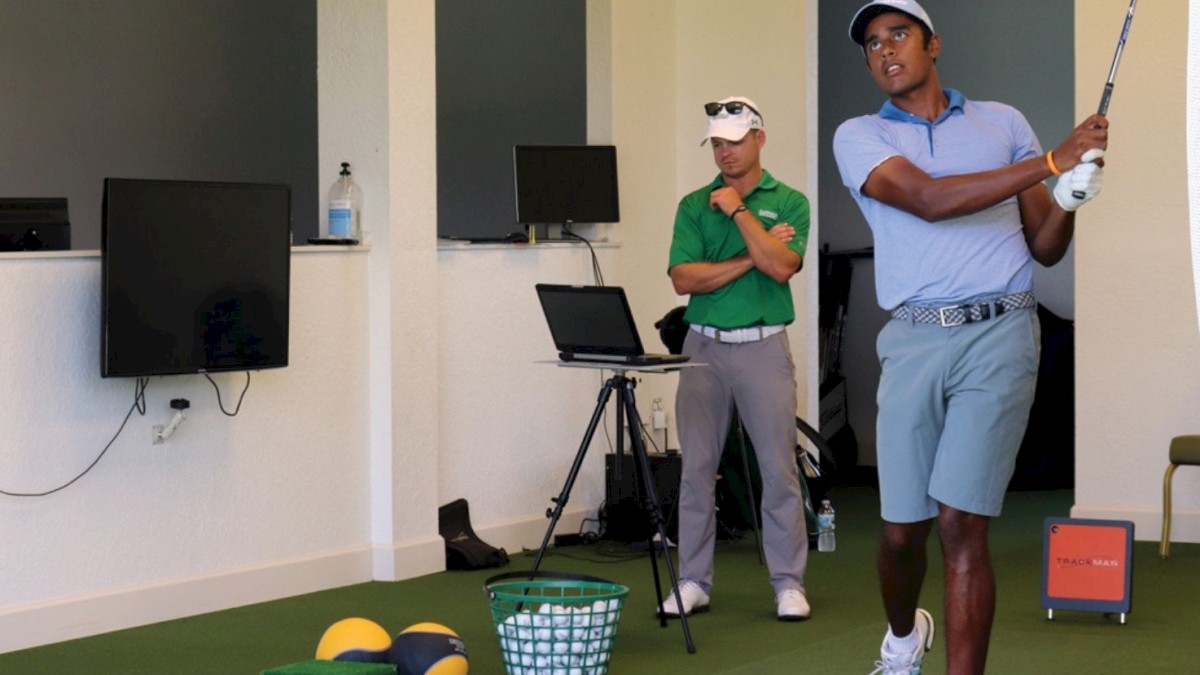 A man in golf attire swings a club in an indoor training area with golf balls and equipment. Another man watches intently.