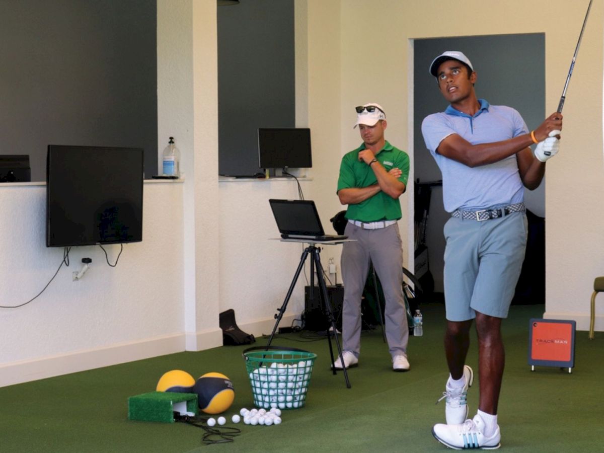 A man in golf attire swings a club in an indoor training area with golf balls and equipment. Another man watches intently.