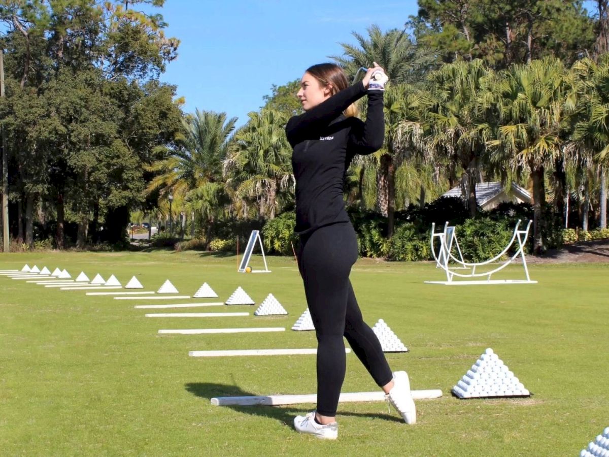 A person dressed in black is practicing their golf swing on a driving range with white golf ball pyramids and targets in the background.