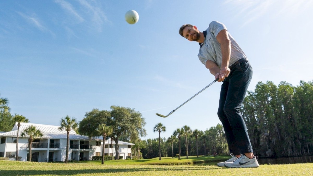 A man is playing golf on a sunny day, hitting a golf ball mid-air with a club, with a white house and trees in the background.