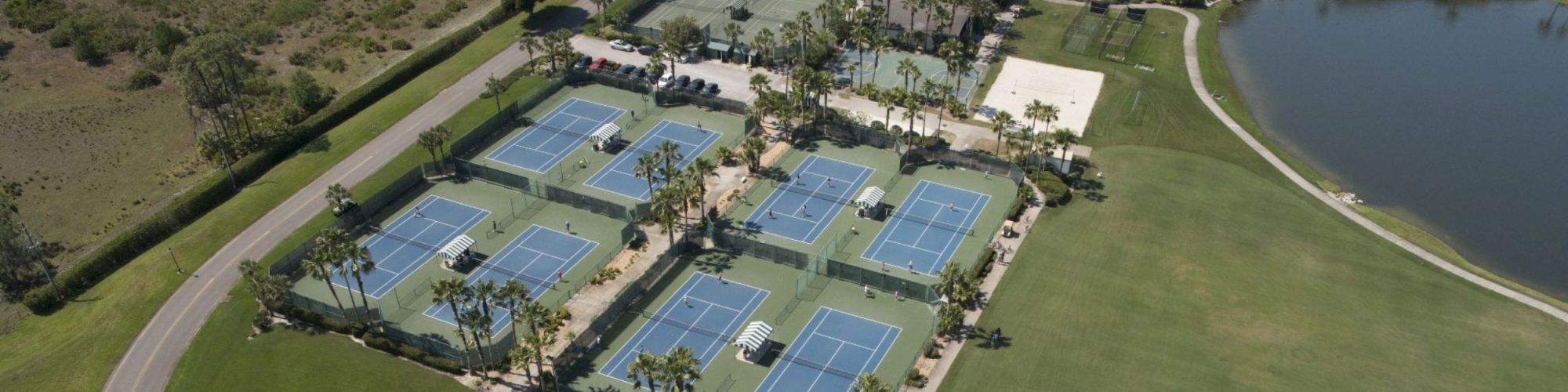 Aerial view of a complex with several tennis courts, surrounding greenery, a large pool, and an adjacent lake.