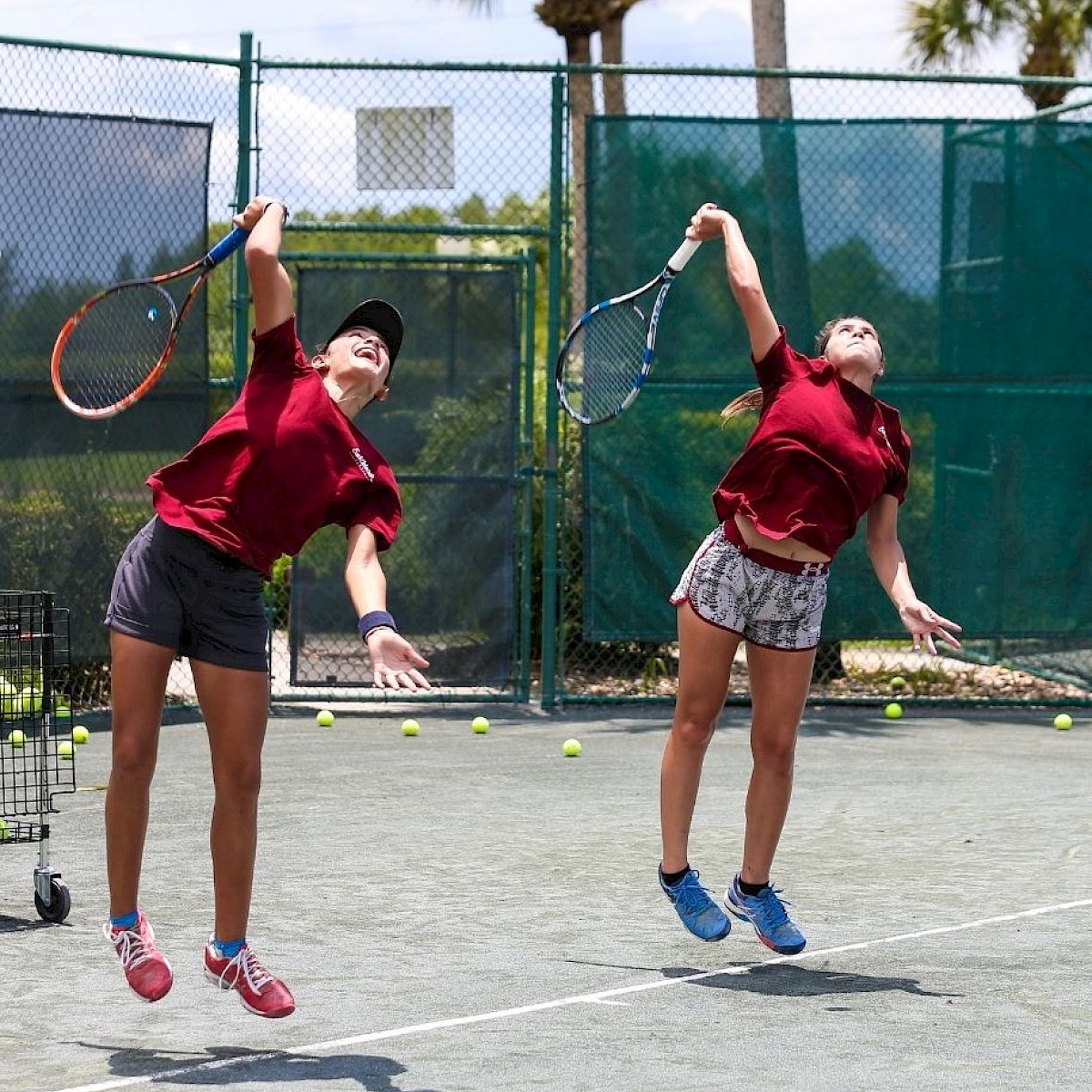 Two people are playing tennis on an outdoor court, both mid-jump, about to hit the ball. They are wearing red shirts and shorts, with tennis rackets in hand.