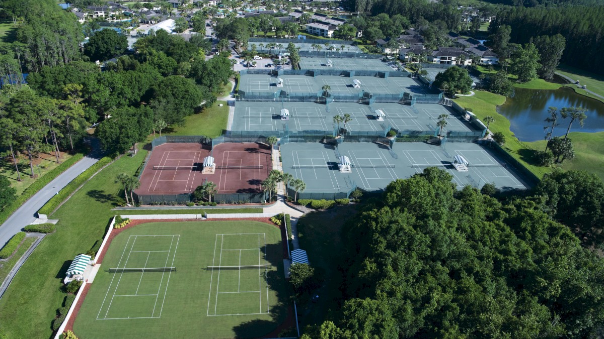 An aerial view of a sports complex with multiple tennis courts, including grass, clay, and hard courts, surrounded by trees and a small pond.