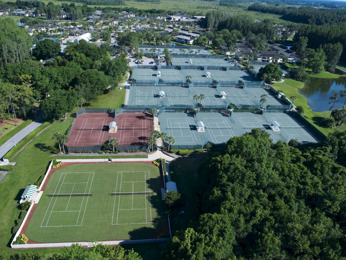 An aerial view of a sports complex with multiple tennis courts, including grass, clay, and hard courts, surrounded by trees and a small pond.