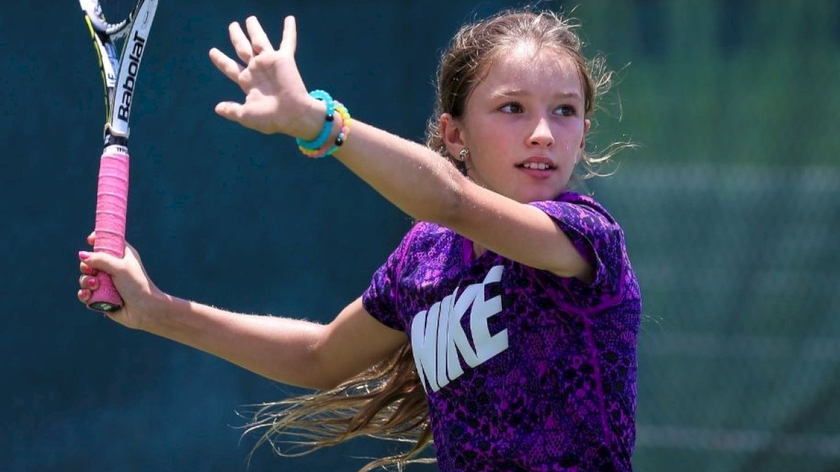 A young girl is playing tennis, holding a racket and mid-swing with a focused expression, wearing a purple shirt with 