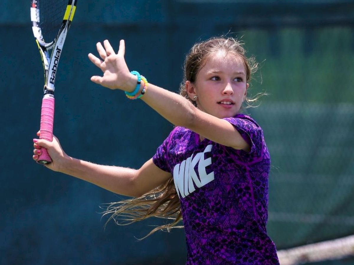 A young girl is playing tennis, holding a racket and mid-swing with a focused expression, wearing a purple shirt with 