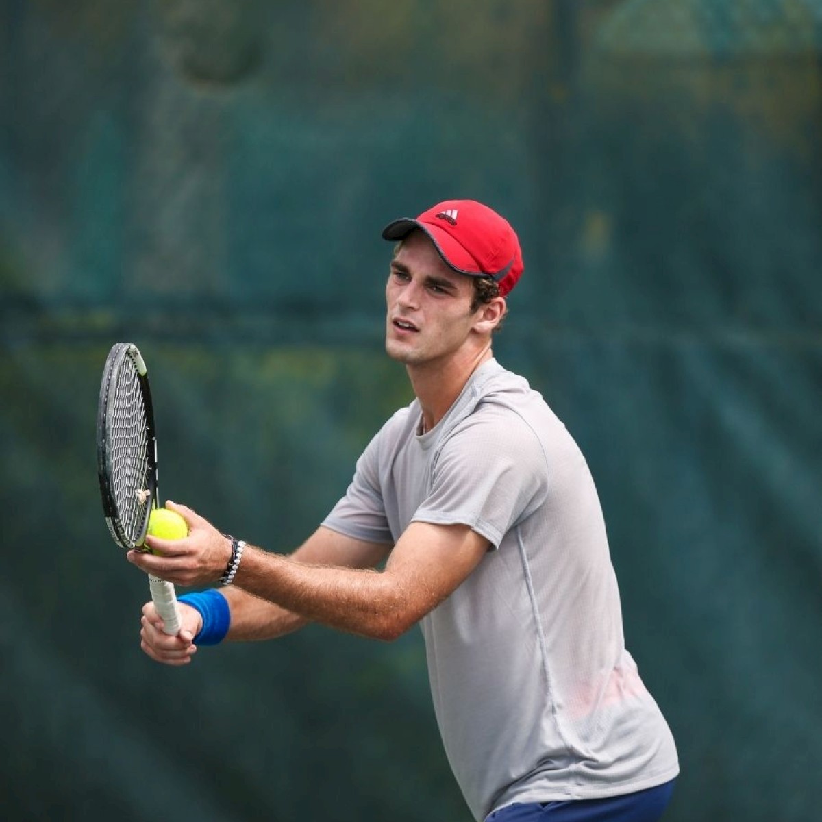 A person in a red cap and grey shirt is holding a tennis racket and preparing to serve a tennis ball on a court with a green background.