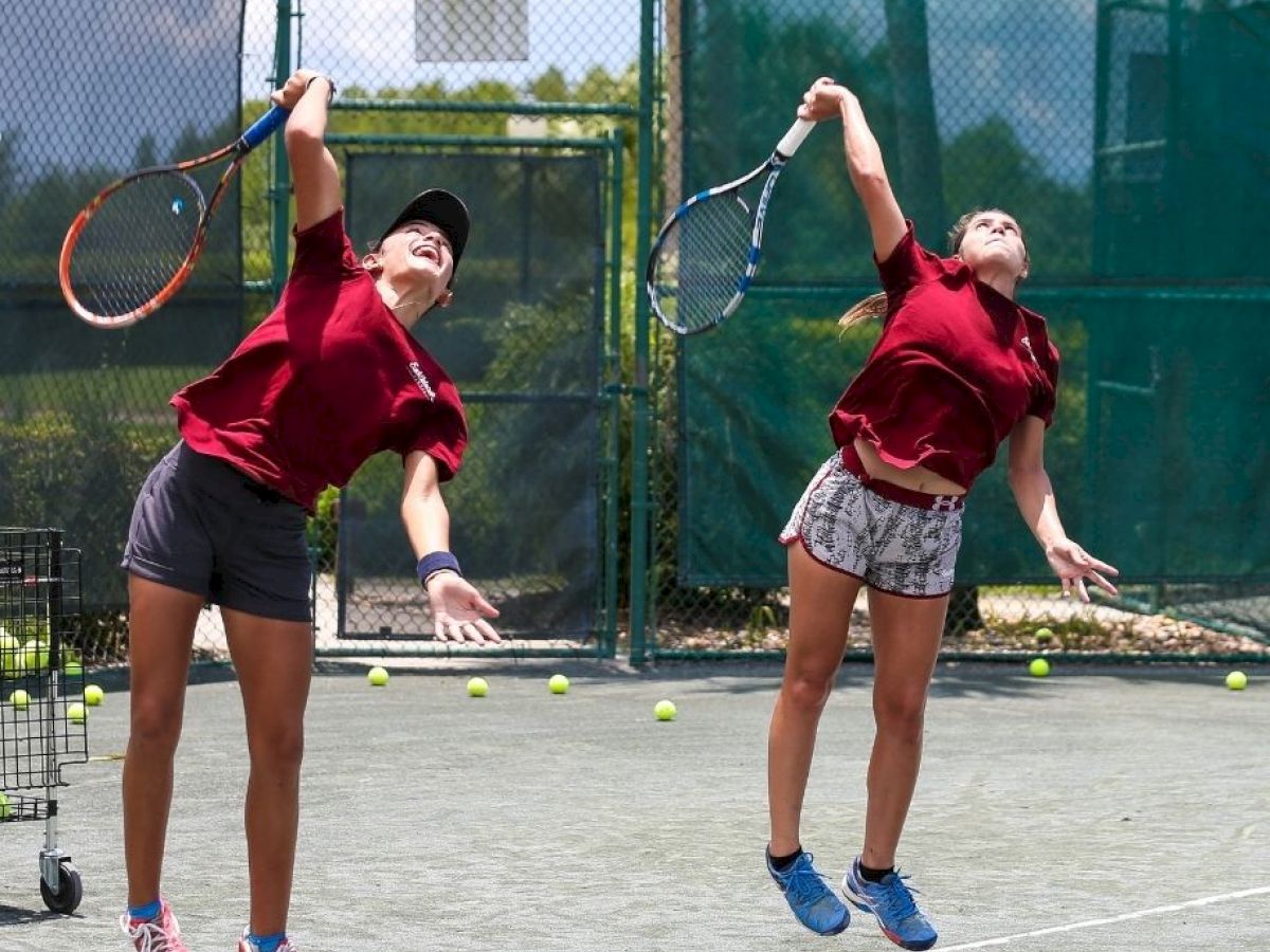 Two young tennis players jump in the air to hit the ball on a sunny outdoor court, surrounded by tennis balls and a ball cart.