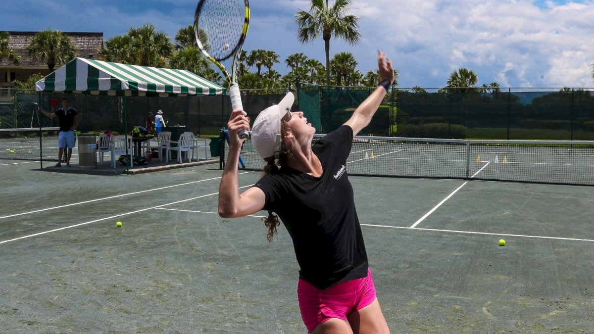 A tennis player in pink shorts and a black shirt prepares to serve on an outdoor court, with people and greenery in the background.