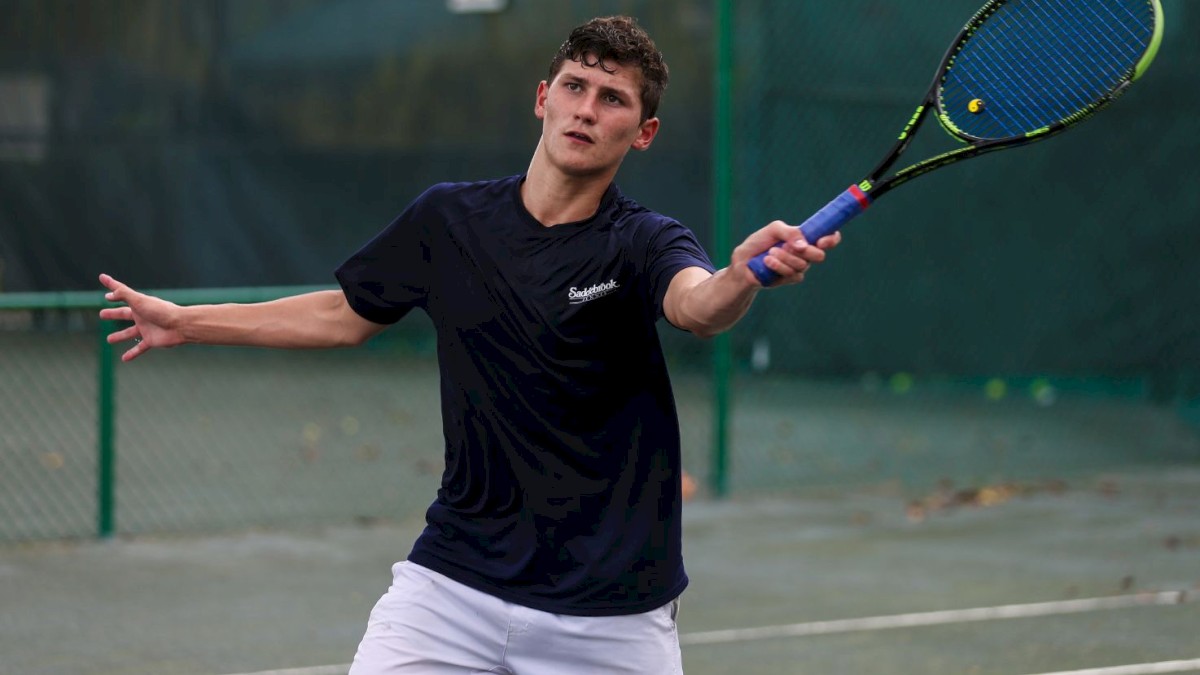 A person is playing tennis, reaching up to hit the ball with a racket on an outdoor court, wearing a black shirt and white shorts.