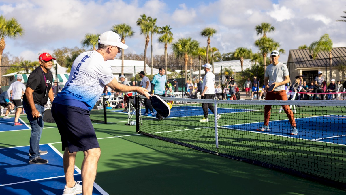 People are playing pickleball on outdoor courts surrounded by palm trees, with spectators and other players in the background.