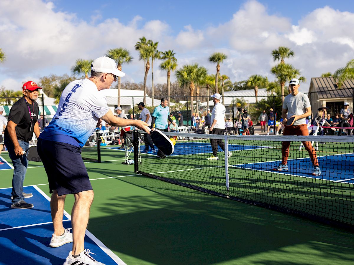 People are playing pickleball on outdoor courts surrounded by palm trees, with spectators and other players in the background.
