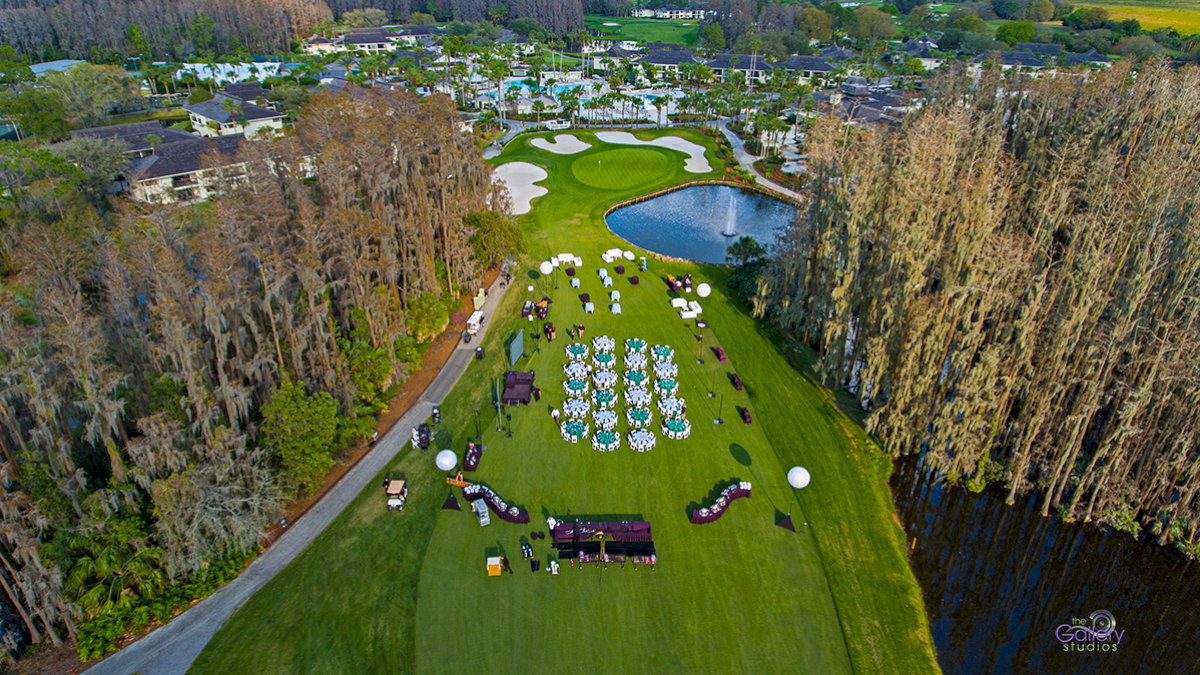 Aerial view of a lush golf course setup for an outdoor event with rows of chairs, tables, and decorations, adjacent to a pond and lined with trees.