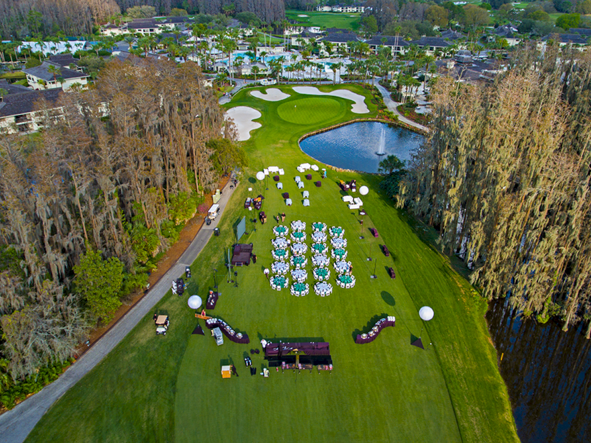 Aerial view of a lush golf course setup for an outdoor event with rows of chairs, tables, and decorations, adjacent to a pond and lined with trees.