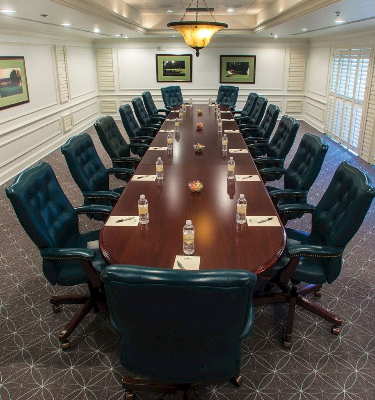 A boardroom with a long table, green chairs, water bottles, and notepads set up for a meeting in a room with framed artwork and a chandelier.