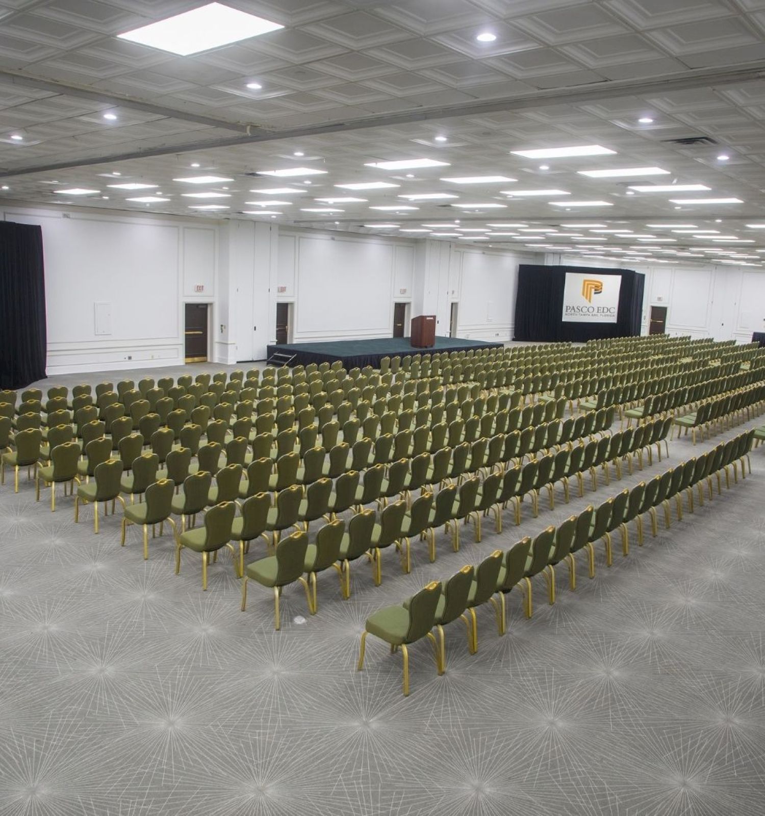A large conference room set up with multiple rows of green chairs facing two projection screens displaying 