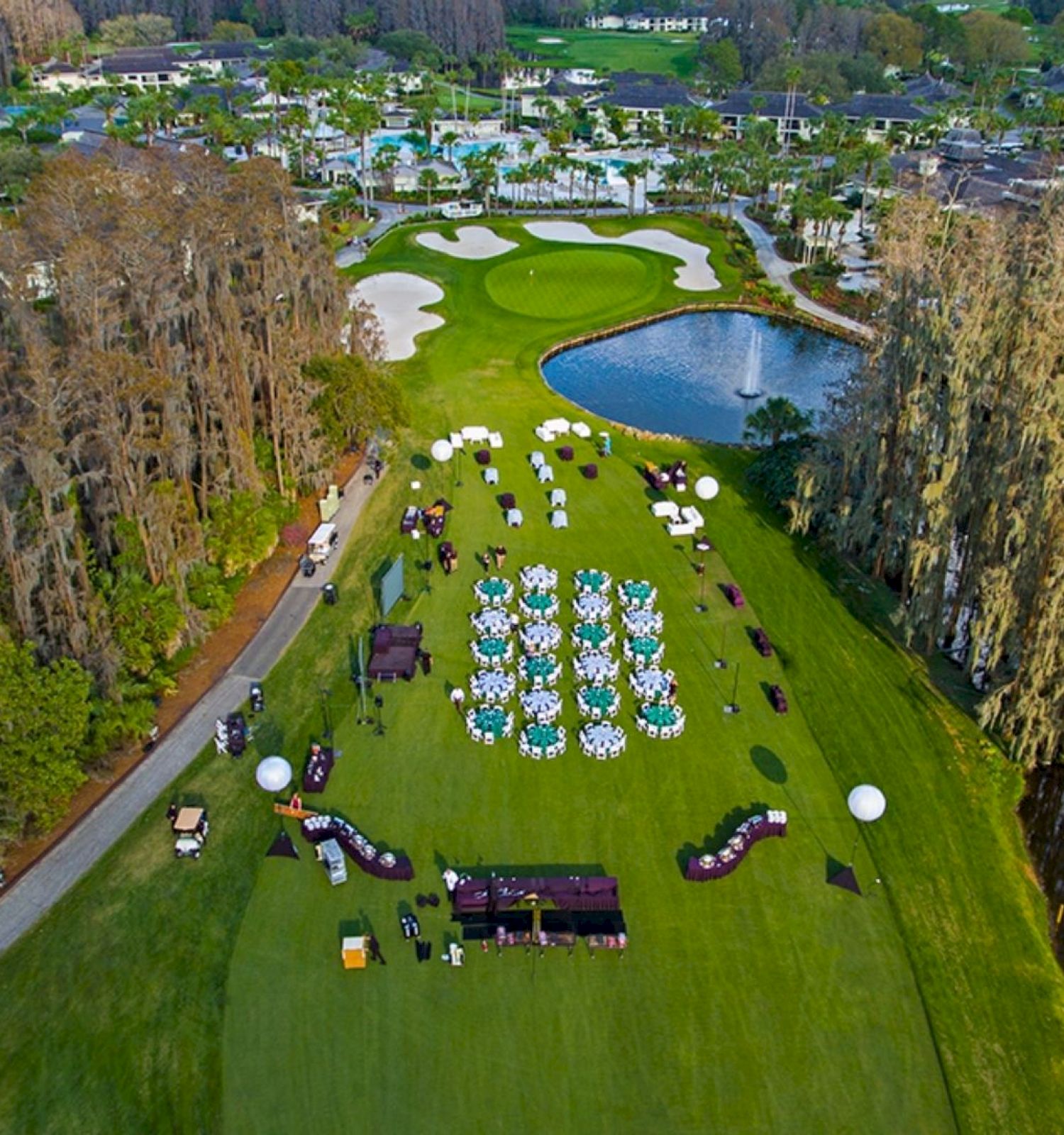 An aerial view shows an event setup on a golf course with tables and chairs, alongside a pond and surrounded by lush greenery and trees.