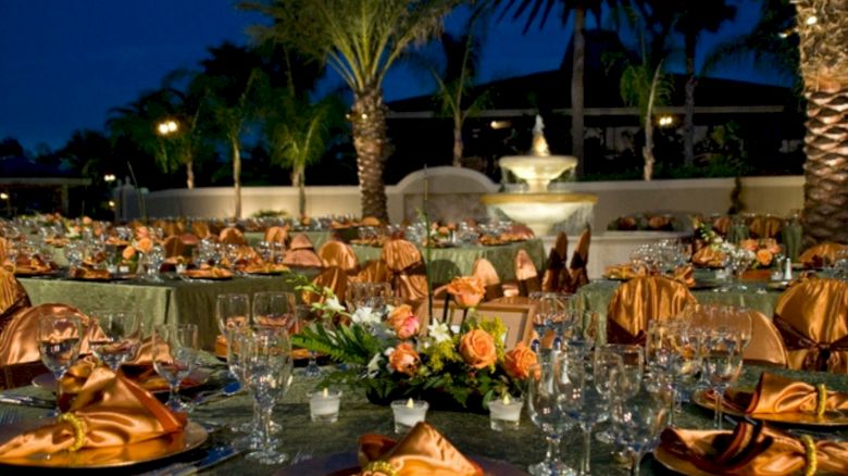 An elegant outdoor event set up with decorated tables, glassware, and napkins; palm trees and a fountain are visible in the background.
