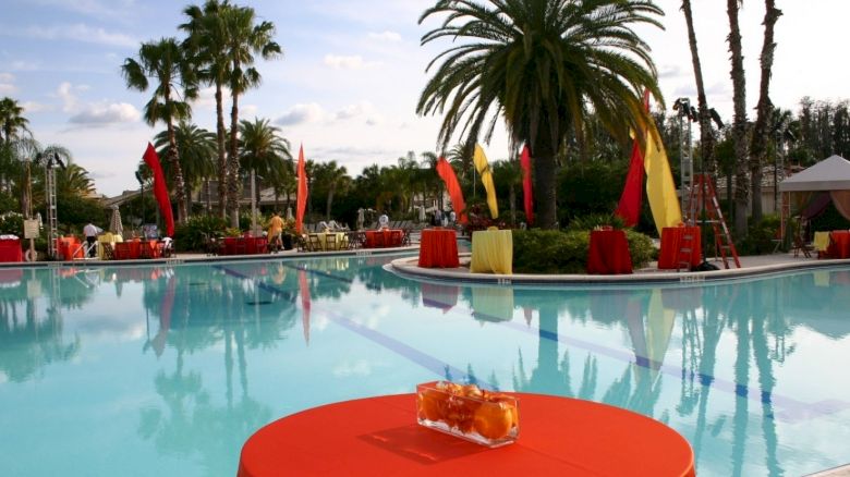 A swimming pool surrounded by palm trees and colorful decorations, with a table covered in an orange cloth and a centerpiece in the foreground.