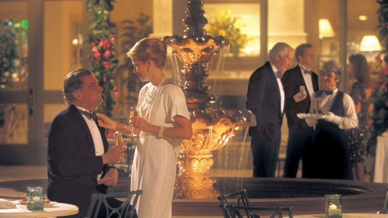 A formal gathering with people in evening wear near a fountain, engaging in conversation and holding drinks and food, in a well-lit area.