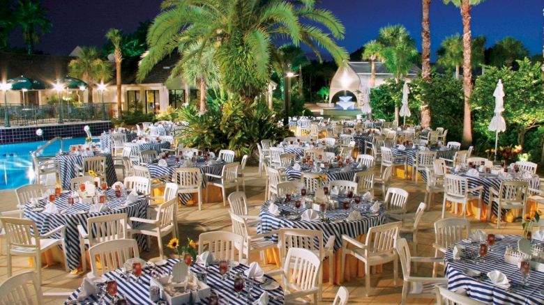 An outdoor dining area by a pool, adorned with round tables covered in striped tablecloths, surrounded by chairs, set up for an event or party.