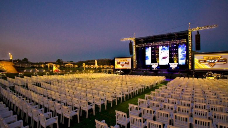 An outdoor event setup with rows of white chairs facing a large stage with screens and lighting. The sky is transitioning to dusk, creating an evening ambiance.