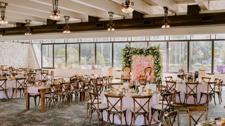 A decorated event space with round tables and wooden chairs arranged for a gathering, featuring a large floral backdrop.